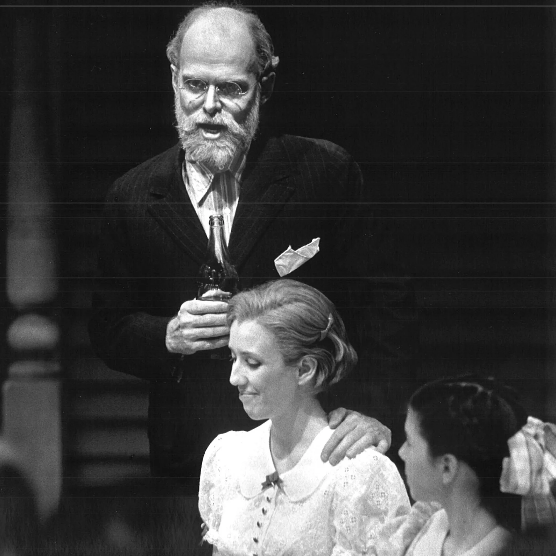 A woman and a girl are sitting at a table together while a man is standing behind them while putting his hand on the woman's shoulder.